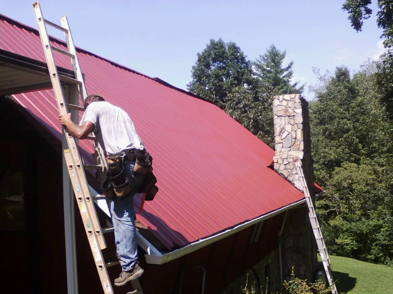 Roofer on a ladder beside a red metal roof and stone chimney — Soffit & Fascia Repair by Roofing Brothers in New Lexington